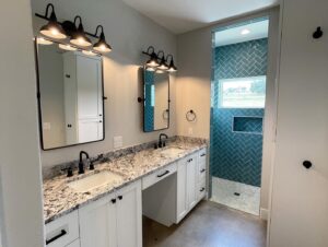 A modern bathroom features a double vanity with granite countertops, two rectangular mirrors, and black light fixtures above each sink. Adjacent is a walk-in shower with blue herringbone tiles, white trim, and a recessed shelf.