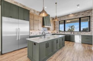 A modern kitchen with green cabinetry, a large stainless steel refrigerator, and a farmhouse sink. Beige brick backsplash, wooden flooring, and two pendant lights hanging above a spacious island with a built-in sink complete the elegant and functional design.