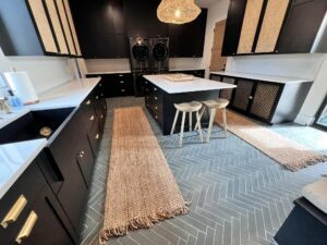 A modern kitchen with dark cabinetry and gold handles, featuring a large island with two beige stools. The floor has a herringbone tile pattern with two woven beige rugs. A woven pendant light hangs above the island, and the countertops are white.