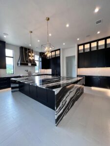 A modern kitchen features black cabinetry, a large black and white marble island, and two geometric pendant lights hanging above. The space is illuminated by large windows and recessed ceiling lights, with sleek high-end appliances and a spacious layout.