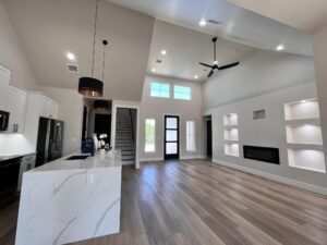 A modern living space with a high ceiling, large windows, and hardwood flooring. It features a white kitchen with black appliances and a marble countertop island, a black ceiling fan, recessed lighting, a fireplace, and built-in wall shelves. Stairs lead to an upper level.