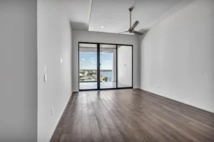 A spacious, empty room with light gray walls, a ceiling fan, and dark wood flooring. The room features large sliding glass doors leading to a balcony with a scenic view of a waterfront and clear blue sky.