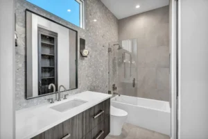 Modern bathroom with a sleek vanity, integrated sink, and a wall-mounted rectangular mirror. A glass-enclosed bathtub and shower combination is on the right, adjacent to light grey tiled walls. The room is well-lit, featuring a small window near the ceiling.