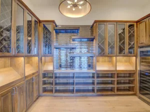 A modern wine cellar with wooden cabinetry and glass-fronted storage units. A central display area features diamond-shaped wine racks. The room is well-lit by a contemporary ceiling light fixture, and the floor is made of light-colored wood.