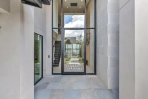 A modern entrance hall with large glass doors and windows, revealing a view into the house and through to the backyard. The interior features sleek, light-colored stone walls and flooring, with a staircase to the left and a pathway outside.