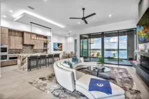 Modern open-plan living area featuring a sleek kitchen with a long island and bar stools, a white circular sofa with a blue throw, a wall-mounted TV, and large sliding glass doors opening to a patio with a scenic lakeside view.