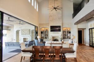 A spacious living room with high ceilings and a stone fireplace. Large windows and sliding glass doors lead to an outdoor patio. The room features a wooden dining table and chairs, gray couches, built-in shelving around the fireplace, and modern decor elements.